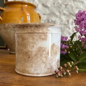 Peut inclure: Un pot en céramique cylindrique présentant un émail blanc avec des taches brunes marbrées d'aspect vieilli et une texture craquelée visible sur toute la surface. Le récipient a un design simple à parois droites avec un bord légèrement relevé.