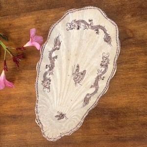 May include: A cream-colored ceramic dish shaped like a large seashell with a scalloped edge. The surface features dark brown illustrations of three dragons and two butterflies arranged around the center, with a decorative brown border.