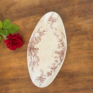 May include: An oval-shaped ceramic tray with a cream-coloured base and a brown transferware pattern featuring floral vines, hanging geometric frames, and candle holders. A single deep red rose with green leaves is positioned beside the tray.