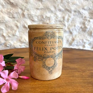 May include: A cylindrical beige stoneware jar with a dark brown printed label reading Confitures Fines Felix Potin Paris, featuring an ornate decorative border and a circular emblem, accompanied by pink oleander flowers on the side.
