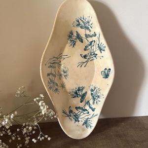 Könnte beinhalten: Eine längliche, mandelförmige Keramikschale mit cremefarbener Glasur. Die Innenfläche zeigt einen blauen botanischen Druck von Geißblattblüten und einem einzelnen Schmetterling, mit einem feinen Craquelé-Finish auf der gesamten Glasur.