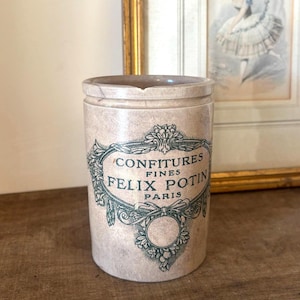 May include: A cylindrical beige stoneware jar featuring a dark green printed label that reads CONFITURES FINES FELIX POTIN PARIS, surrounded by an ornate floral and ribbon border. The jar has a smooth finish and a small chip on the top rim.