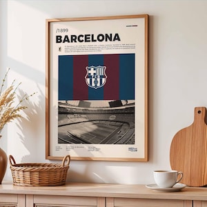 May include: A framed poster featuring the FC Barcelona crest and stadium. The poster has a cream-colored background with the text "BARCELONA" and other details. The crest is centered above a blue and maroon striped design.