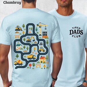 Op de afbeelding: Lichtblauwe T-shirts. Eén shirt heeft een bouwthema-afbeelding met wegen, voertuigen en gebouwen. Het andere shirt heeft de tekst "TIRED DADS CLUB" in zwart. Beide shirts zijn korte mouwen.