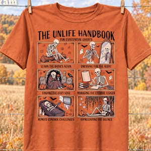 Op de afbeelding: Een oranje t-shirt met een afbeelding getiteld THE UNLIFE HANDBOOK FOR EXISTENTIAL GHOSTS. De print bevat zes panelen met skeletten die verschillende activiteiten uitvoeren met de bijschriften: LEARN THE BASICS AGAIN, DRESSING FOR THE AUDIT, FINDING THE COZY VOID, MANAGING THE ETERNAL LEDGER, REMOTE CONTROL CHALLENGES en APPRECIATING THE SILENCE.