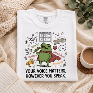 May include: A white t-shirt featuring a green frog wearing a red cape and holding a microphone. The graphic includes a communication board, a keyboard, a megaphone, and the text: "SUPPORT", "EMPATHY", "UNDERSTAND", "INCLUSION", "YOUR VOICE MATTERS, HOWEVER YOU SPEAK."