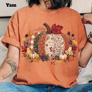 Puede incluir: Una camiseta naranja de manga corta con un gran gráfico central de una calabaza compuesta por varios segmentos de tela estampada. La calabaza está rodeada de hojas de otoño, bayas y calabazas pequeñas. El texto Yam está impreso en negro.
