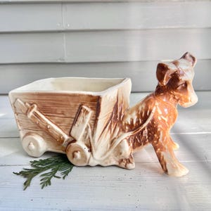 May include: A ceramic dog pulling a brown wagon planter. The dog is brown and cream-colored, with a detailed texture. The wagon is rectangular with a light brown finish and cream-colored wheels. A sprig of greenery rests in front.