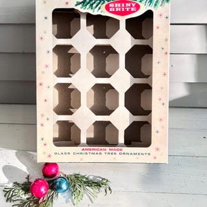 May include: Vintage "Shiny Brite" Christmas ornament box with interior compartments. The box is cream-colored with a green and red logo and text. The box is decorated with starbursts and contains three colorful ornaments.