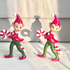 May include: Two ceramic Christmas elf figurines. Each elf is wearing a red hat and jacket, green pants, and red shoes. They are holding a red and white striped candy cane. The elves have a cheerful expression.