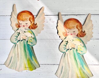 May include: Two vintage paper angel ornaments with wings, holding a white lamb in their arms. The angels are wearing green dresses with white trim and have red hair. The background is a white wooden surface.
