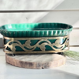 May include: A green ceramic planter with a decorative brass stand. The stand features a floral design and is perfect for displaying plants or flowers.