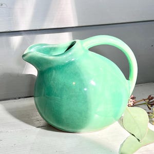 May include: A small, round, mint-green ceramic pitcher with a curved handle and a spout. The pitcher is glossy and sits on a white surface. The background is a light gray wall with shadows.