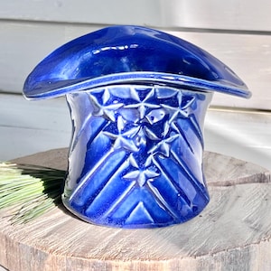 May include: A blue ceramic planter shaped like a hat with a wide brim and a geometric pattern of stars. The planter is glazed in a deep blue color.