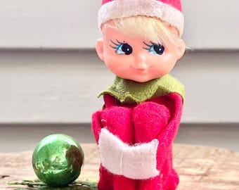 May include: A small elf figurine with blonde hair, blue eyes, and long eyelashes, wearing a red felt suit with a white-trimmed hat and a green collar. The elf is sitting with knees pulled up, holding a small white book, next to a green ornament.