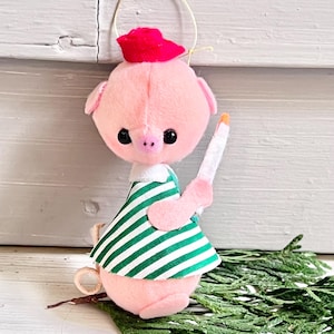 May include: A pink pig ornament with a red hat and a green and white striped dress. The pig is holding a white candle with an orange flame. A gold wire loop is attached to the top for hanging. The ornament is approximately 4 inches tall.