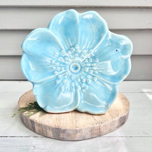 May include: A light blue ceramic flower-shaped dish with a detailed center and petal design. The dish sits on a round, natural wood slice, showcasing a rustic aesthetic. The flower dish is a decorative item.