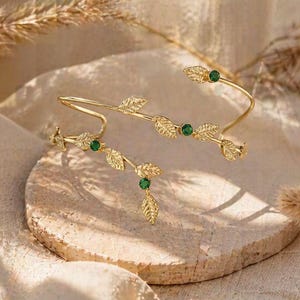May include: A gold-colored arm cuff and matching bracelet, both featuring a leaf design with small green gemstones. The jewelry is displayed on a textured, light-colored surface, with dried plants in the background. The cuff is approximately 7 inches in length.