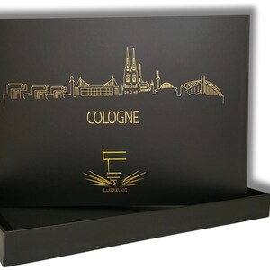 May include: Black box with a gold skyline of Cologne, Germany, and the city's name in gold lettering. The box also features a logo with the text "LASERKUNST". The box is open, revealing a tray.