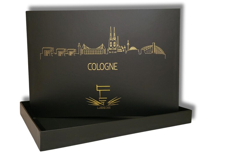 May include: Black box with a gold skyline of Cologne, Germany, and the city's name in gold lettering. The box also features a logo with the text "LASERKUNST". The box is open, revealing a tray.