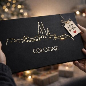 May include: Black gift box with a gold skyline of Cologne and the city's name. A tag reads "A Gift for You" with a red heart. The box is held by hands, with a blurred background of Christmas lights.