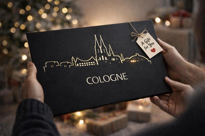 May include: Black gift box with a gold skyline of Cologne and the city's name. A tag reads "A Gift for You" with a red heart. The box is held by hands, with a blurred background of Christmas lights.