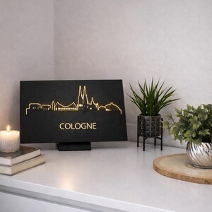 May include: Black rectangular light fixture with the word "COLOGNE" and a gold outline of the Cologne skyline. A candle, books, and plants are on a white surface.