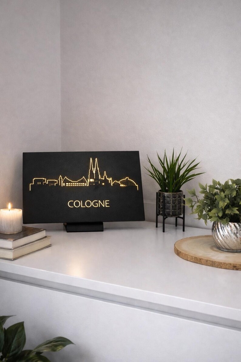 May include: Black rectangular light fixture with the word "COLOGNE" and a gold outline of the Cologne skyline. A candle, books, and plants are on a white surface.