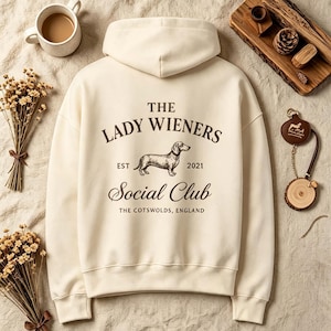 May include: A cream-coloured hooded sweatshirt featuring a dark brown graphic on the back. The text reads THE LADY WIENERS, EST 2021, Social Club, and THE COTSWOLDS, ENGLAND, accompanied by a detailed illustration of a dachshund dog in the centre.