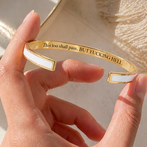 May include: A gold-tone cuff bracelet featuring a white enamel inlay along the outer edges. The interior surface of the metal band is engraved with the black text, This too shall pass, BUT FUCKING HELL, in a clear, serif typeface.