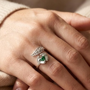 May include: A silver ring featuring a wing design and tiny footprints. The ring has a heart-shaped green gemstone at the center. The ring is on a person's finger.