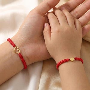 May include: Red braided bracelets with gold heart charms. One bracelet is on an adult's wrist, and the other is on a child's wrist. The bracelets are a matching set, symbolising a bond between two people.