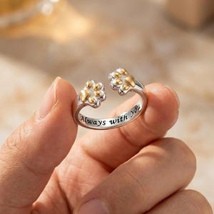 May include: Silver-toned ring with two paw prints, each with gold-colored pads. The ring is open-ended, with the paw prints at each end. The words "Always with You" are engraved on the band. The ring is held in a hand.