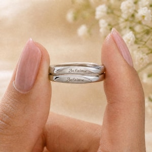 May include: Two silver rings stacked together, each engraved with the phrase "Je t'aime". The rings are held against a soft, blurred background with white flowers. The rings are a simple band design.