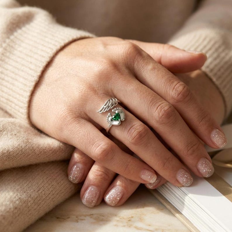 May include: A silver ring featuring a wing design and tiny footprints. The ring has a heart-shaped green gemstone at the center. The ring is on a person's finger.