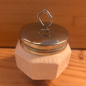 May include: A small, white, hexagonal ceramic jar featuring embossed scrollwork patterns on each side. The container is topped with a polished, reflective silver-tone metal lid that includes a diamond-shaped handle loop at the very center.