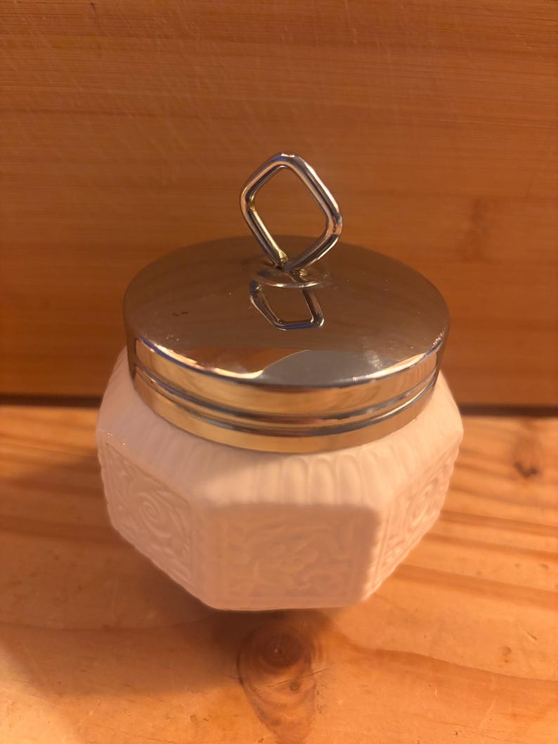 May include: A small, white, hexagonal ceramic jar featuring embossed scrollwork patterns on each side. The container is topped with a polished, reflective silver-tone metal lid that includes a diamond-shaped handle loop at the very center.