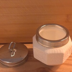 May include: A white octagonal jar with a textured floral pattern on its sides and a metallic silver rim. Beside the jar sits a matching circular metallic silver lid featuring a small vertical loop handle on its top surface for easy removal.