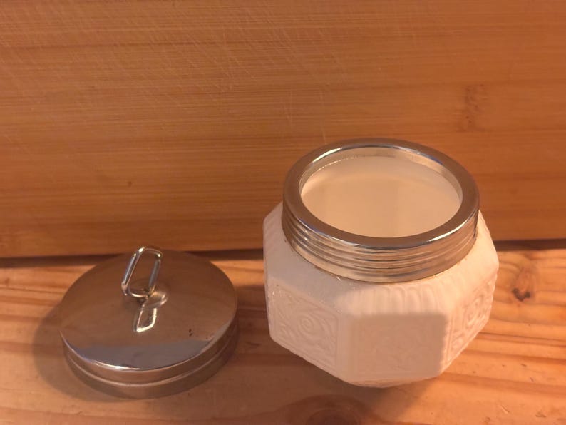May include: A white octagonal jar with a textured floral pattern on its sides and a metallic silver rim. Beside the jar sits a matching circular metallic silver lid featuring a small vertical loop handle on its top surface for easy removal.