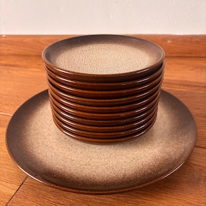 May include: A stack of brown and tan ceramic plates. The plates have a speckled tan center and a dark brown rim. One plate is lying flat beneath the stack. The plates are on a wooden surface.