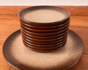 May include: A stack of brown and tan ceramic plates. The plates have a speckled tan center and a dark brown rim. One plate is lying flat beneath the stack. The plates are on a wooden surface.