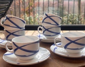 May include: White ceramic teacups and saucers with a blue abstract design. The cups have a rounded shape with a handle, and the saucers are round. The blue design is a series of intersecting lines. The set is arranged on a wooden surface.