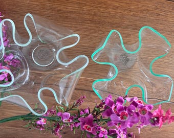 May include: Two clear glass decorative bowls with wavy edges, outlined in light green. The bowls are on a wooden surface, with purple flowers and green stems in the foreground. The bowls have a unique, artistic design.