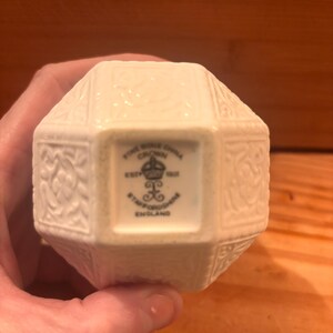 May include: A cream-colored, multi-faceted fine bone china object with an embossed floral pattern. The base features a black stamp reading: FINE BONE CHINA, CROWN, EST 1801, STAFFORDSHIRE, ENGLAND, with a crown and decorative symbol in the center.