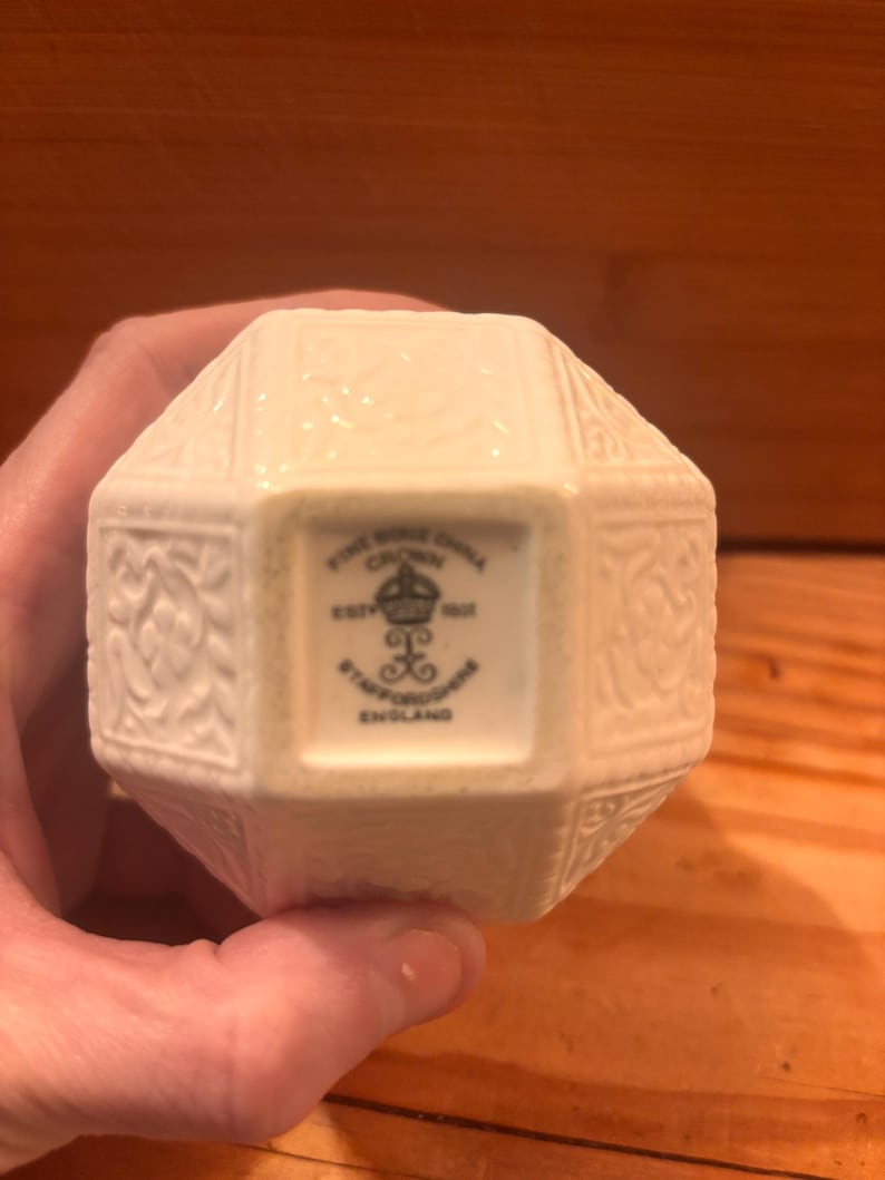 May include: A cream-colored, multi-faceted fine bone china object with an embossed floral pattern. The base features a black stamp reading: FINE BONE CHINA, CROWN, EST 1801, STAFFORDSHIRE, ENGLAND, with a crown and decorative symbol in the center.