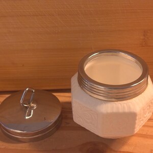 May include: A white ceramic jar with an octagonal body featuring embossed floral patterns and a metallic silver rim. The jar includes a matching circular metallic silver lid with a small vertical loop handle on top, shown detached from the jar.