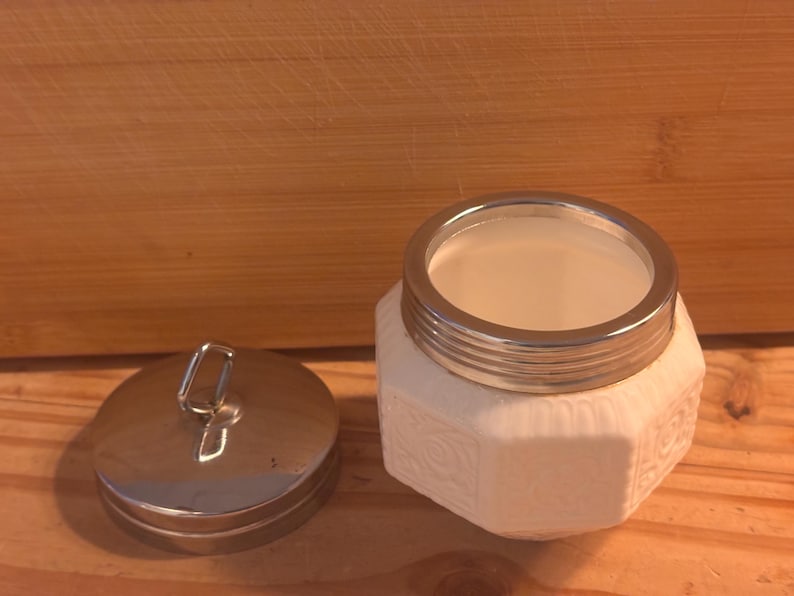 May include: A white ceramic jar with an octagonal body featuring embossed floral patterns and a metallic silver rim. The jar includes a matching circular metallic silver lid with a small vertical loop handle on top, shown detached from the jar.