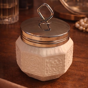May include: A white ceramic jar with an octagonal body featuring embossed floral patterns on each side. The jar is topped with a polished silver-toned metal lid that includes a distinctive diamond-shaped handle centered on the top surface.