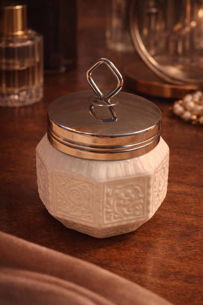 May include: A white ceramic jar with an octagonal body featuring embossed floral patterns on each side. The jar is topped with a polished silver-toned metal lid that includes a distinctive diamond-shaped handle centered on the top surface.
