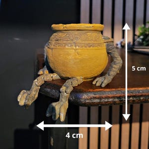 Könnte beinhalten: Ein skurriler, gelb-goldener Keramiktopf mit Armen und Beinen, der auf einer dunklen Holzoberfläche sitzt. Der Topf ist etwa 5 cm hoch und 4 cm breit. Die Arme und Beine sind in einem strukturierten, graubraunen Farbton gehalten, was die einzigartige, handwerkliche Ästhetik unterstreicht.